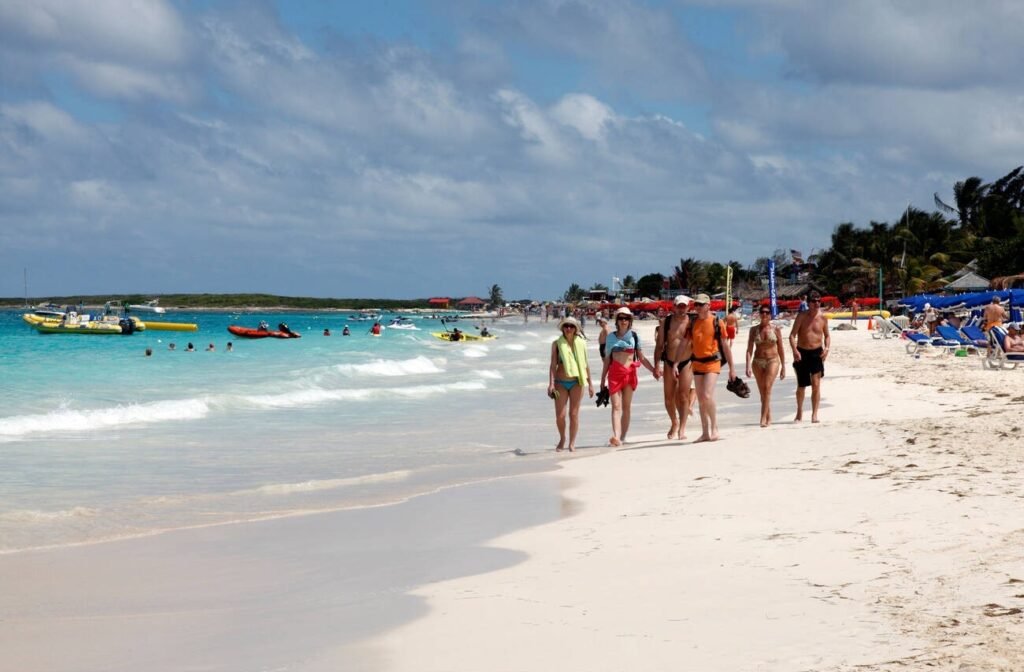 A group of tourists walking along the shoreline of the lively Orient Bay Beach in St. Martin, with turquoise water and water sports in the background.