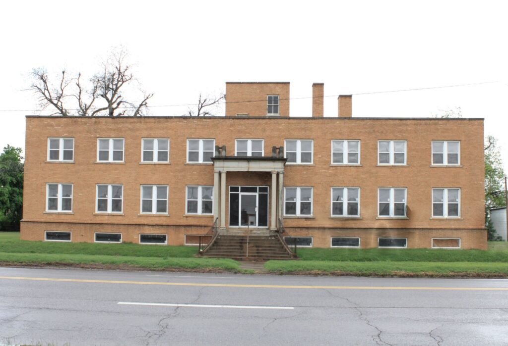 A large, abandoned three-story brick building with two chimneys in Chickasha, Oklahoma, on an overcast day.