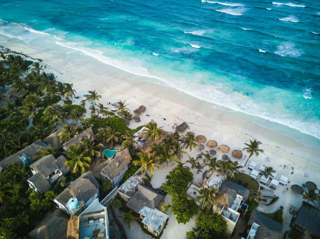 An aerial view of the white sand beaches and turquoise water of Tulum, Mexico, lined with eco-chic resorts and palm trees.