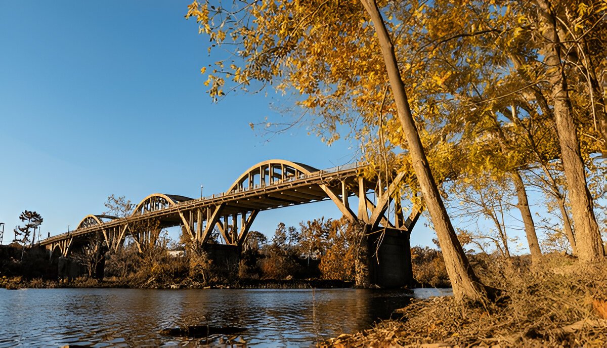 Alabama Towns: The historic Bibb Graves Bridge in Wetumpka, Alabama, seen from the riverbank with golden autumn leaves in the foreground during sunset.