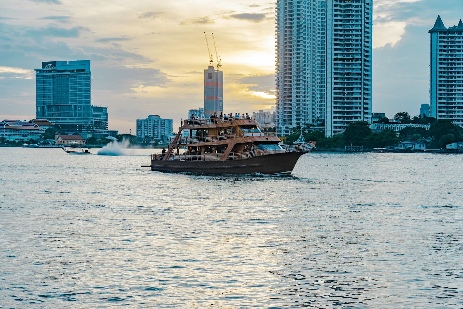 A large multi-deck tourist boat cruises on the Chao Phraya River in Bangkok, with modern high-rise buildings forming the city skyline in the background at dusk.
