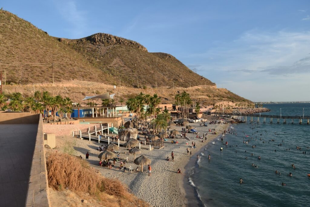 An elevated view of the busy Coromuel Beach in La Paz, Mexico, with desert hills in the background and a pier stretching into the water.