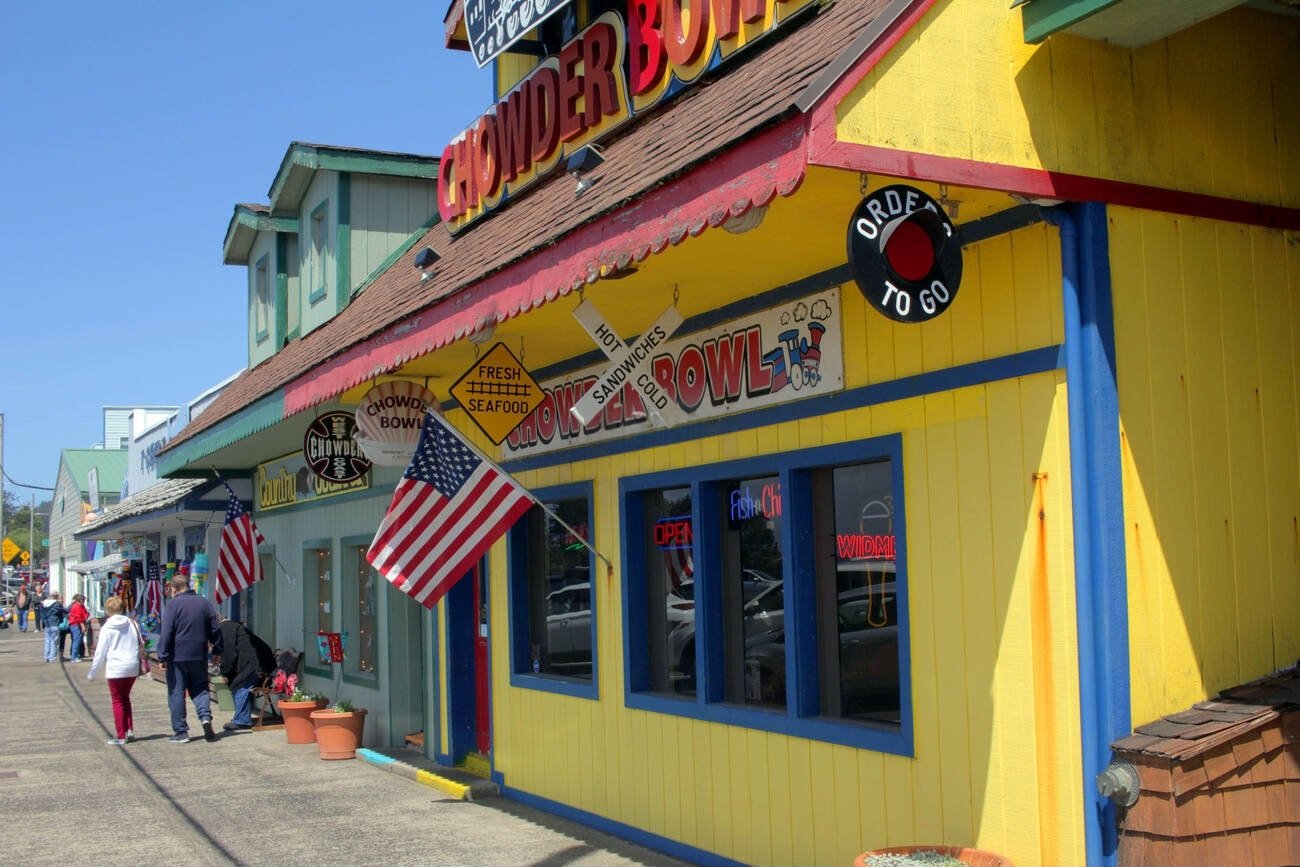 The bright yellow storefront of the Chowder Bowl restaurant in a unique Pacific Coast town, with people walking by on a sunny day.