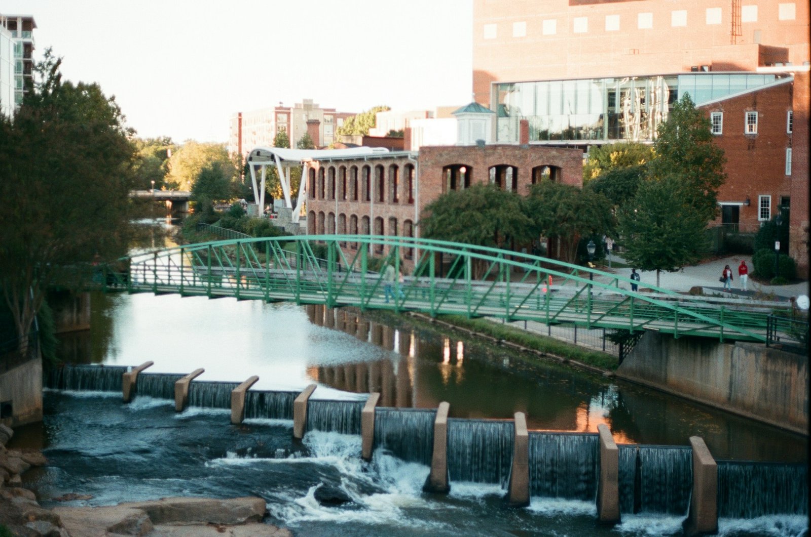 A green pedestrian bridge spans a river with small waterfalls in downtown Greenville, South Carolina, surrounded by brick buildings and trees.
