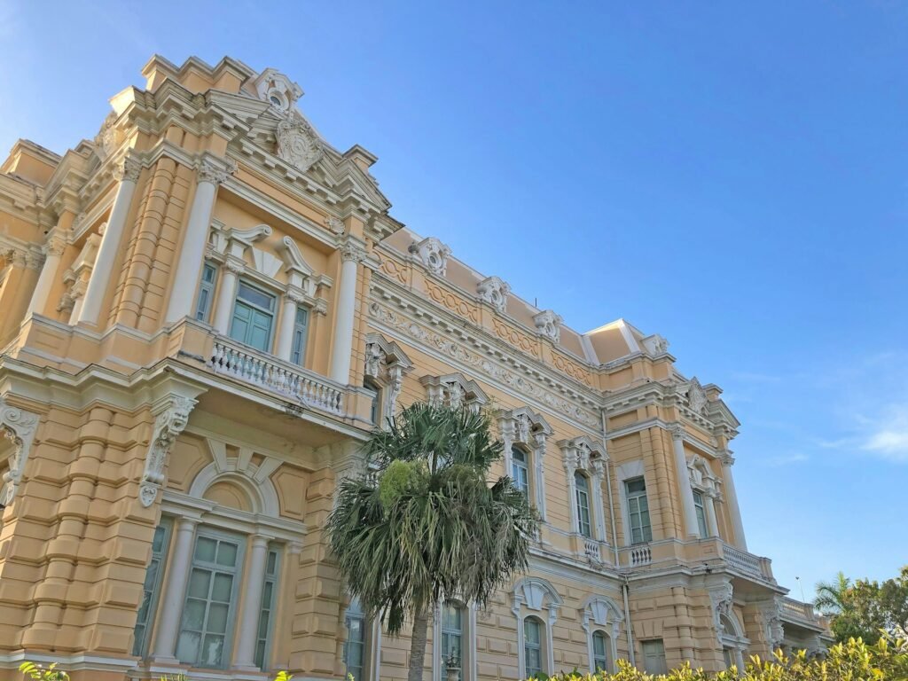 A grand, yellow colonial-era mansion with white trim and a palm tree in front, against a clear blue sky in Mérida, Mexico.