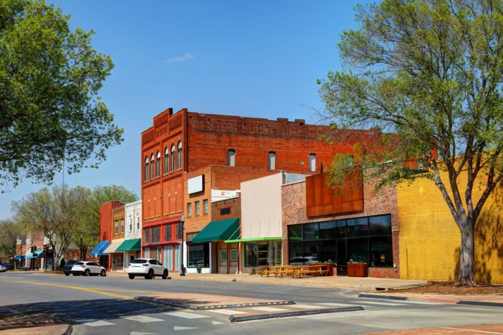 A sunny street scene in historic downtown Stillwater, Oklahoma, showing colorful brick buildings and local shops.
