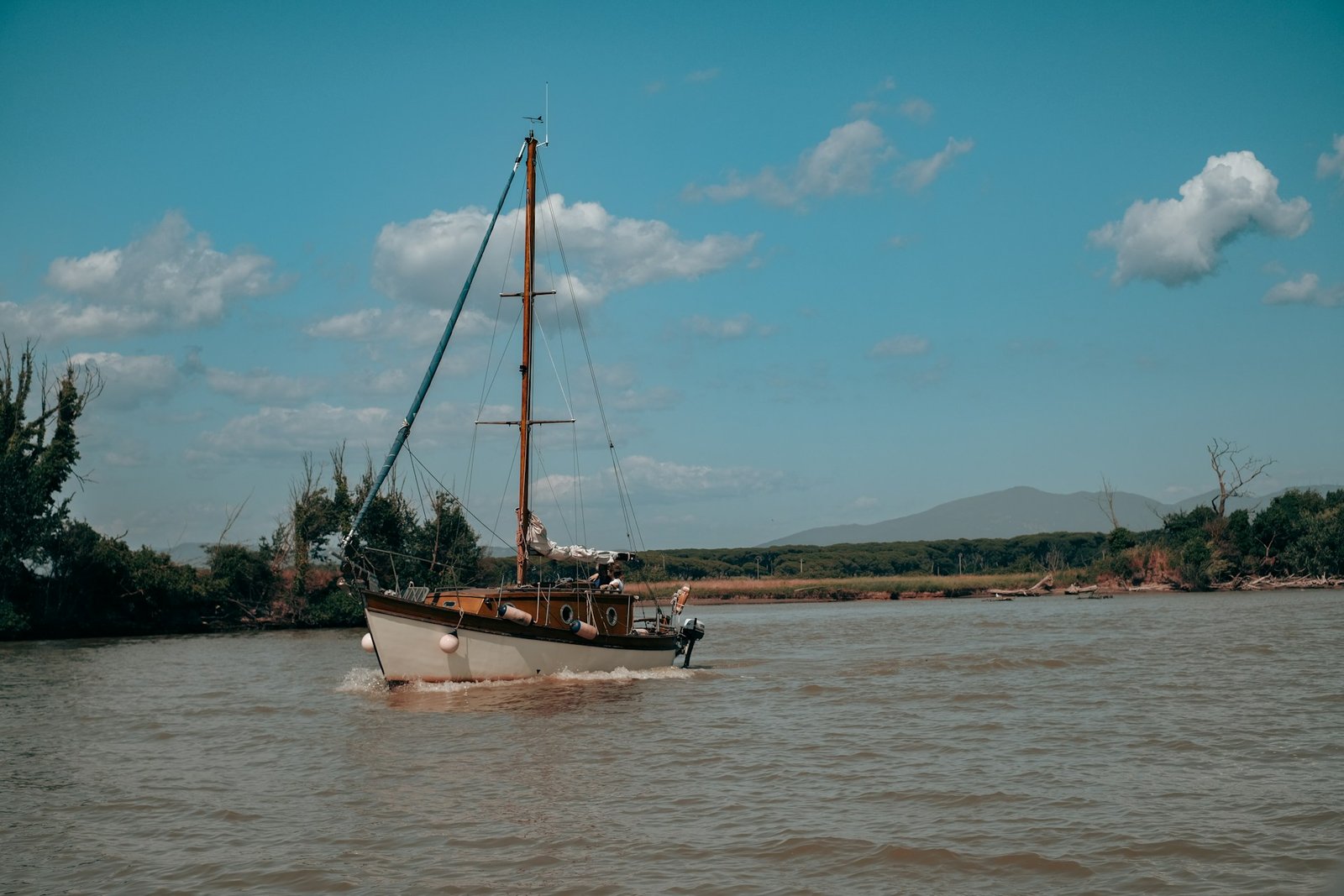 A small white boat with a mast navigates the wide Irrawaddy River, with lush green banks and distant mountains under a blue sky.