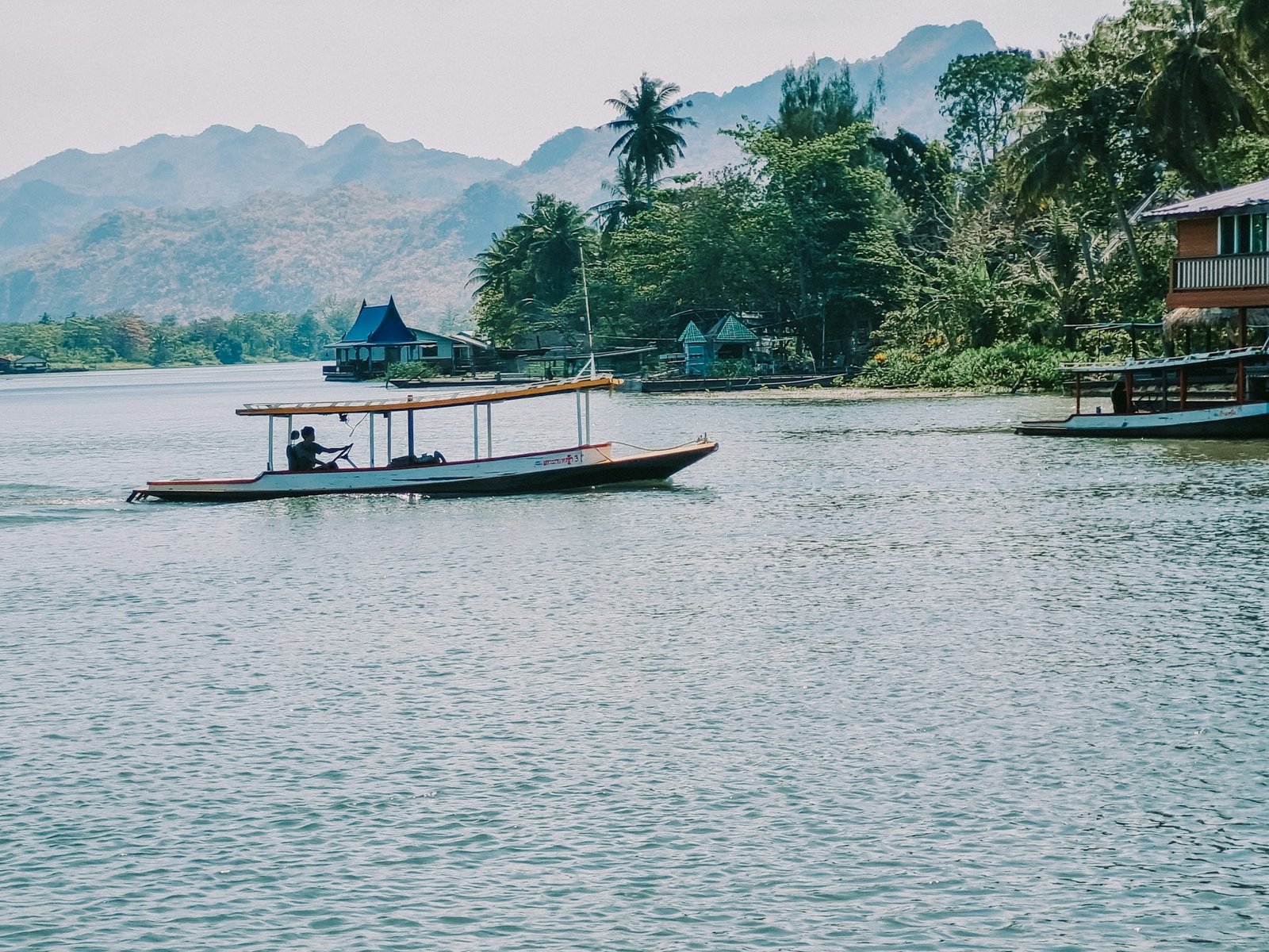 A long, narrow boat with a canopy travels on the Mekong River, framed by lush green mountains and traditional riverside houses.