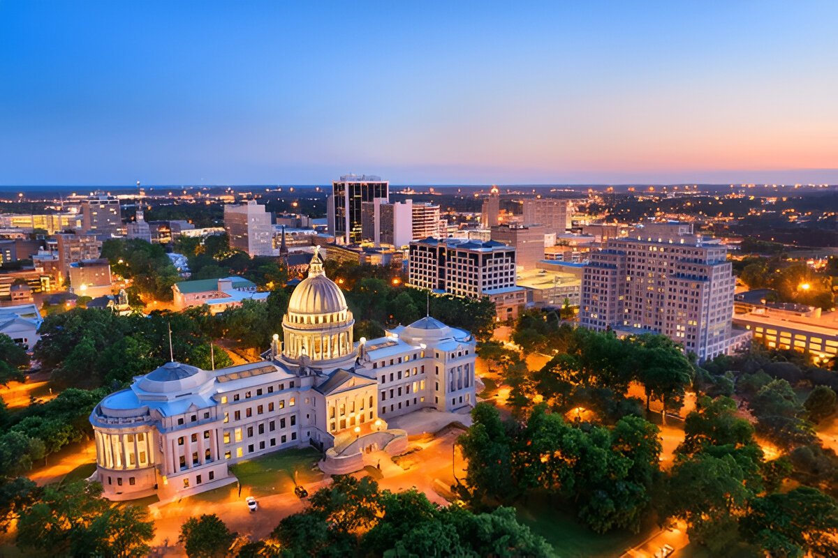 An aerial view of the Mississippi State Capitol building in Jackson, illuminated at dusk with the city skyline in the background.