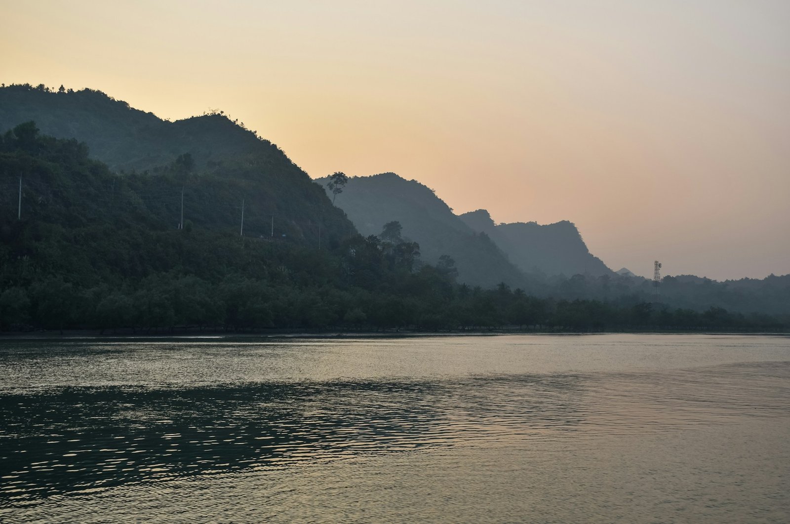 A serene view of the Nam Song River in Laos, with dark, forested limestone mountains rising from the calm water under a hazy sky.