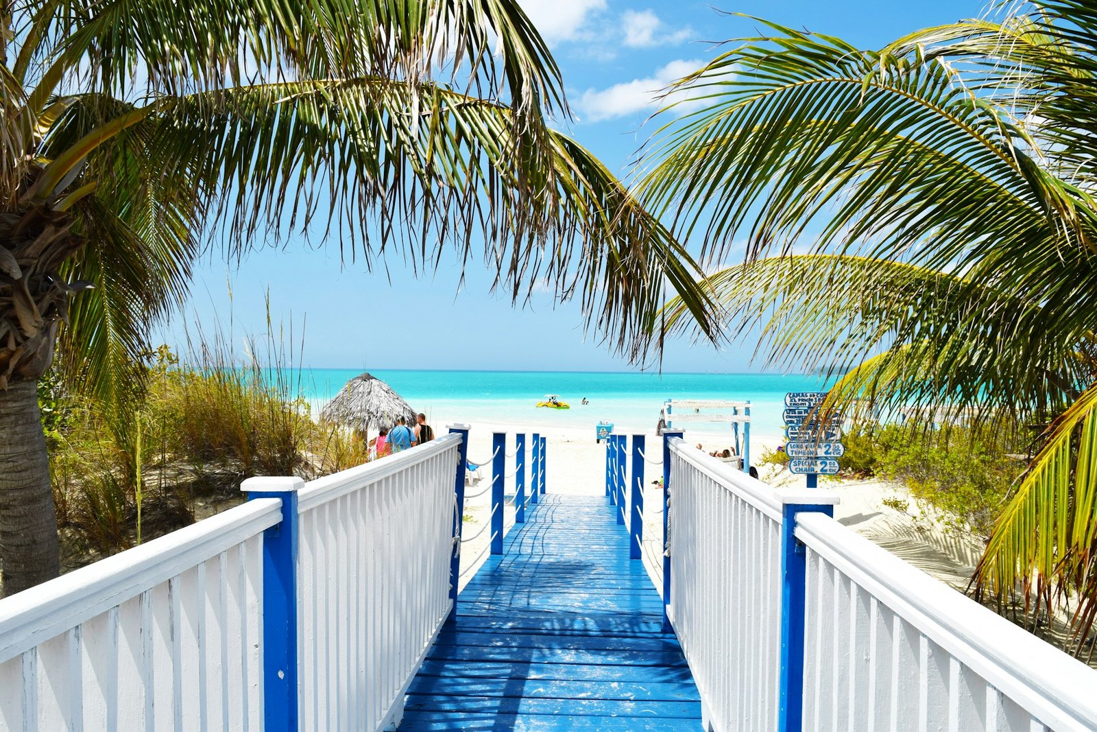 A bright blue wooden walkway leading down to a white sand beach with turquoise water and palm trees in the Caribbean.