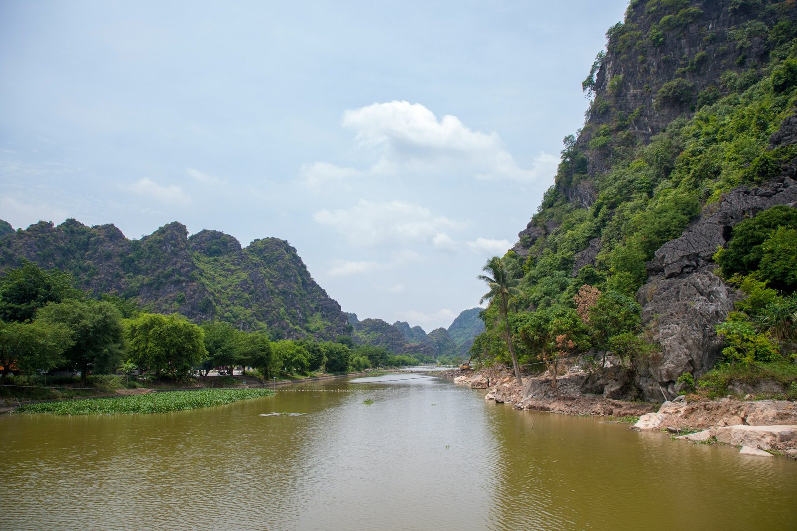 A wide, calm river flows between towering, green-covered karst mountains under a partly cloudy sky, characteristic of Vietnam's Red River region.
