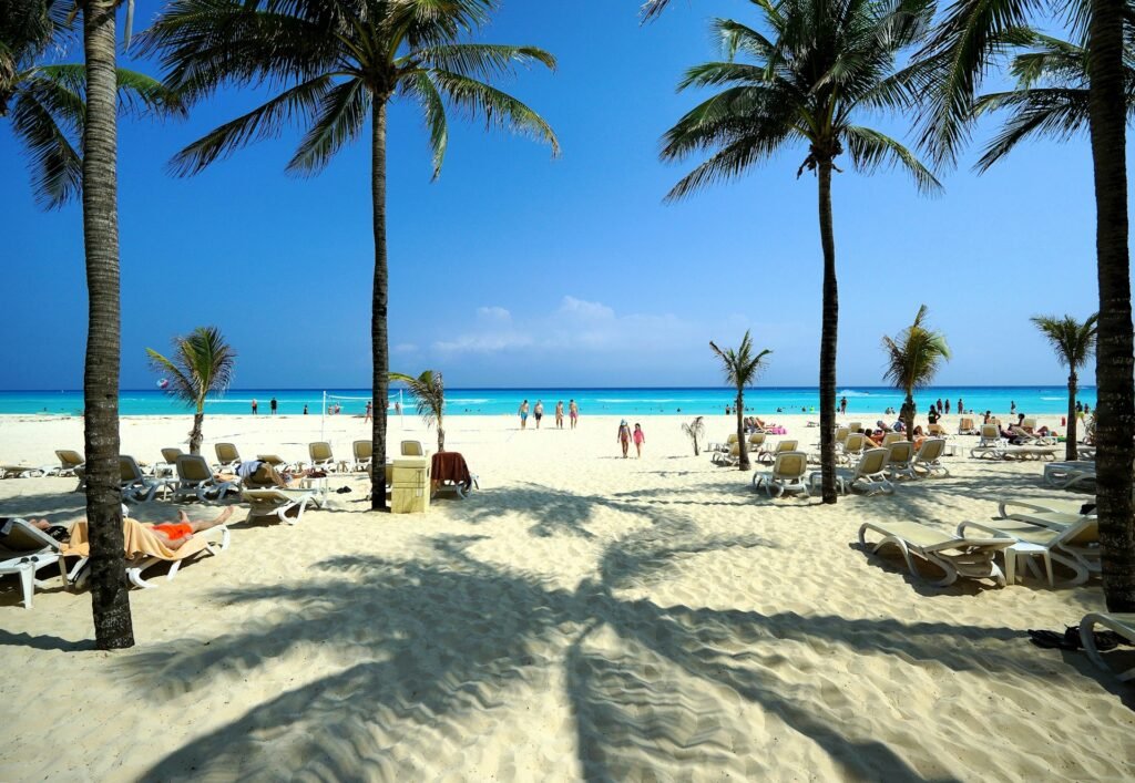 A beautiful white sand beach in Playa del Carmen, Mexico, with palm trees casting shadows and people relaxing by the turquoise ocean.