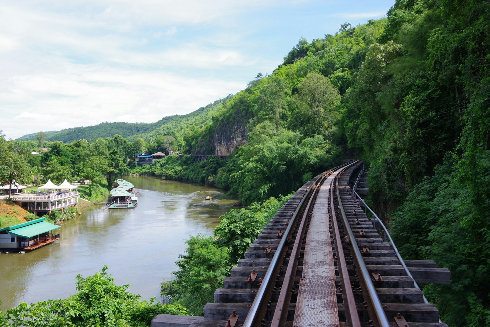 The historic railway track of the Death Railway curves along the River Kwai in Thailand, with lush green forested hills and floating accommodations visible on the river.