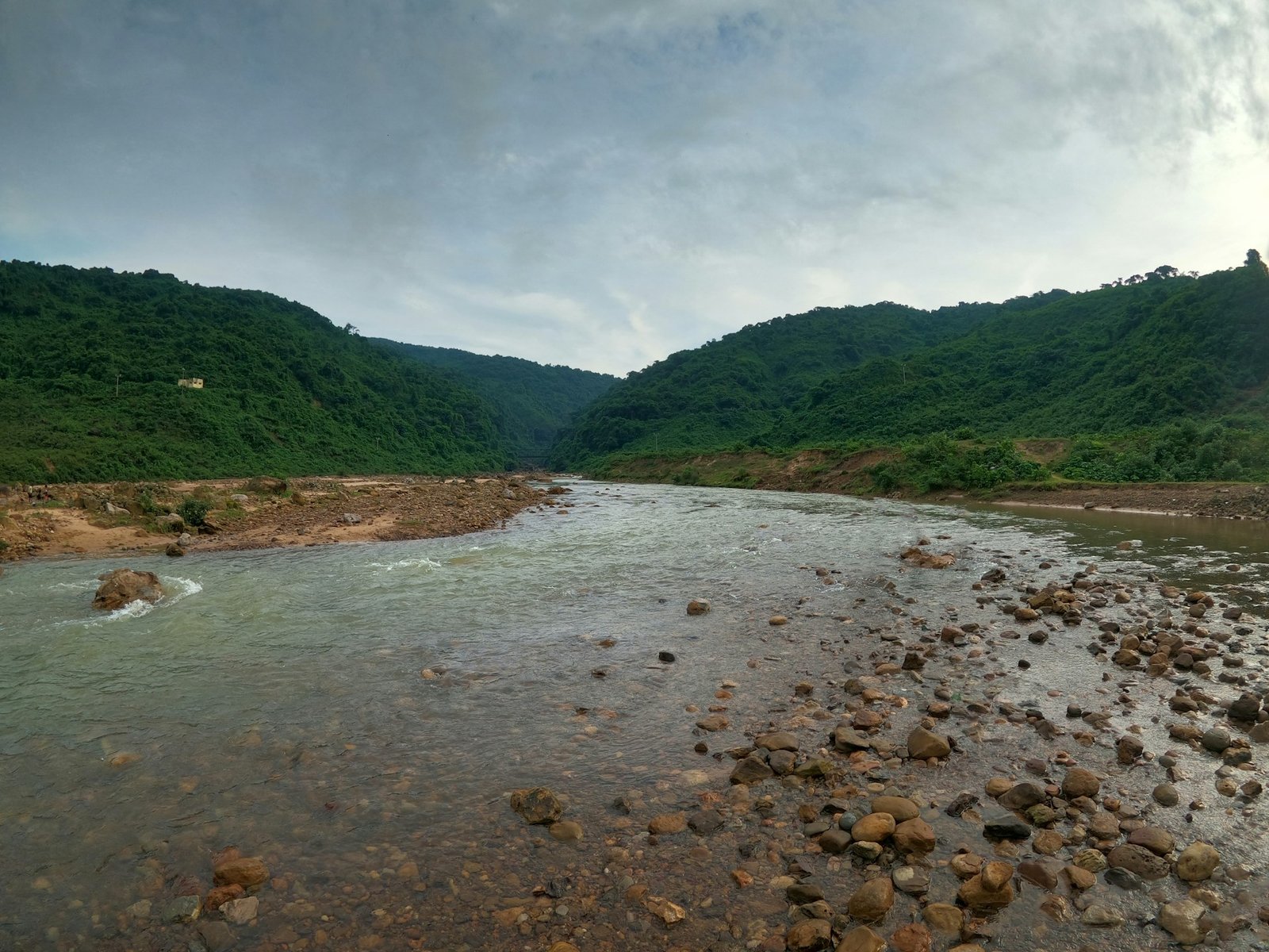 A wide, rocky river flows through a valley between lush green, forested mountains under an overcast sky, representing the remote Salween River.