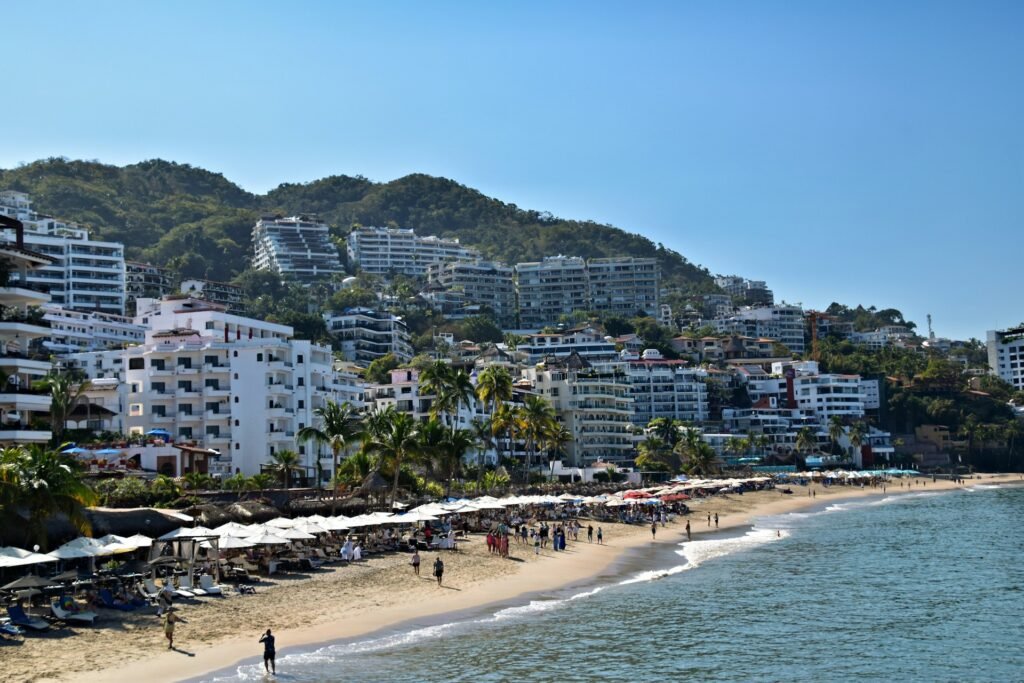 A sunny day at a popular beach in Puerto Vallarta, Mexico, with hotels and resorts built into the green hillsides.