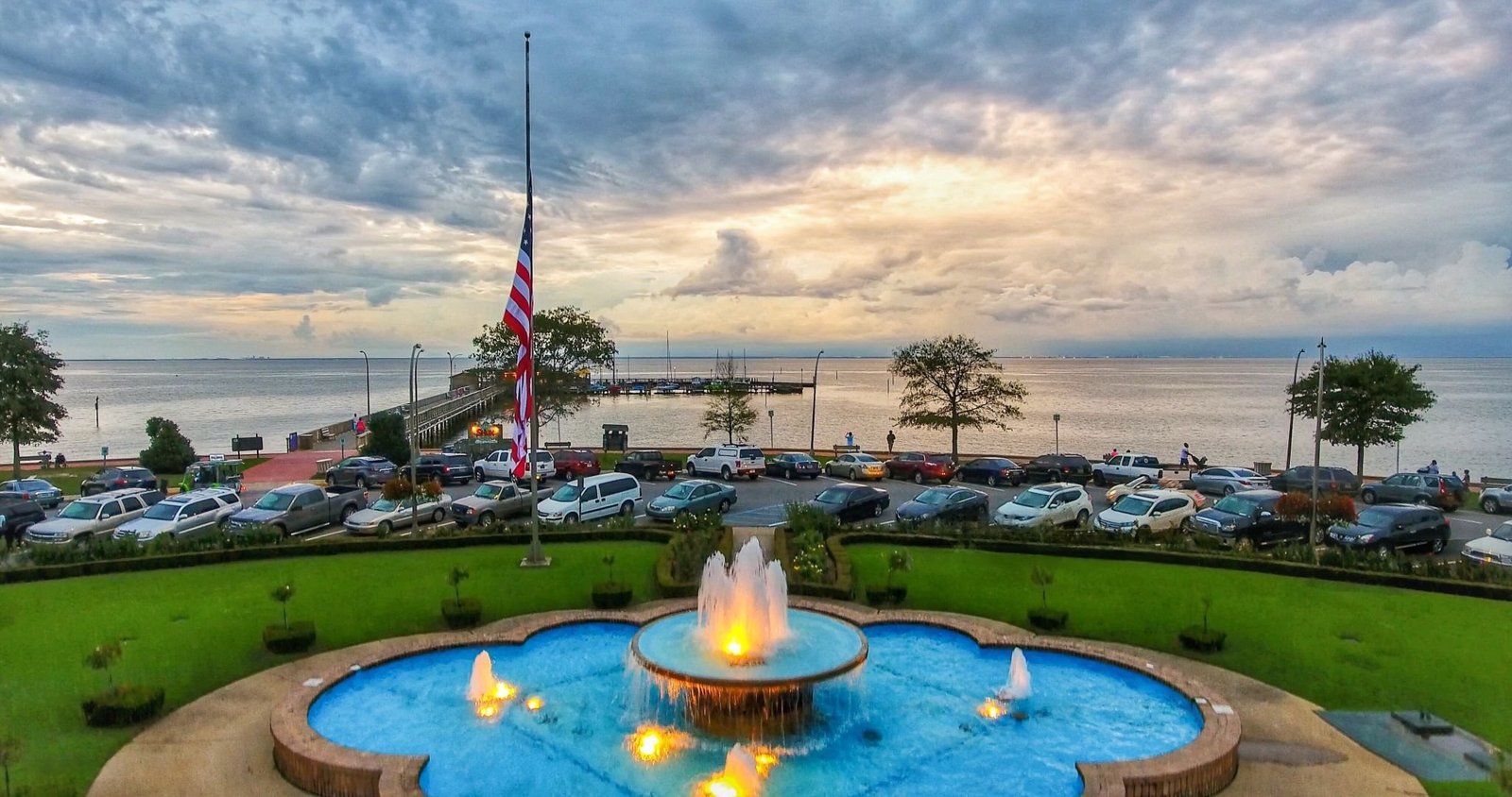 A scenic view of the Fairhope Municipal Pier and Park at sunset, with a large, illuminated fountain in the foreground and Mobile Bay in the background.