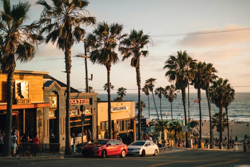 A street scene in a Los Angeles beach town at sunset, with palm trees lining the road and the Pacific Ocean in the background.