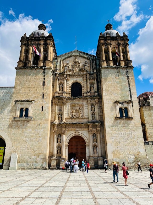 The ornate stone facade of the Templo de Santo Domingo de Guzmán in Oaxaca, Mexico, with tourists in the plaza.