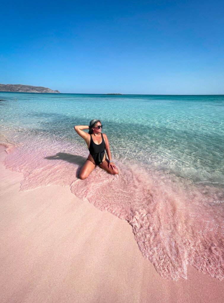 A woman in a black swimsuit kneeling at the water's edge on the famous Pink Sands Beach in the Bahamas, showing the contrast with the turquoise water.