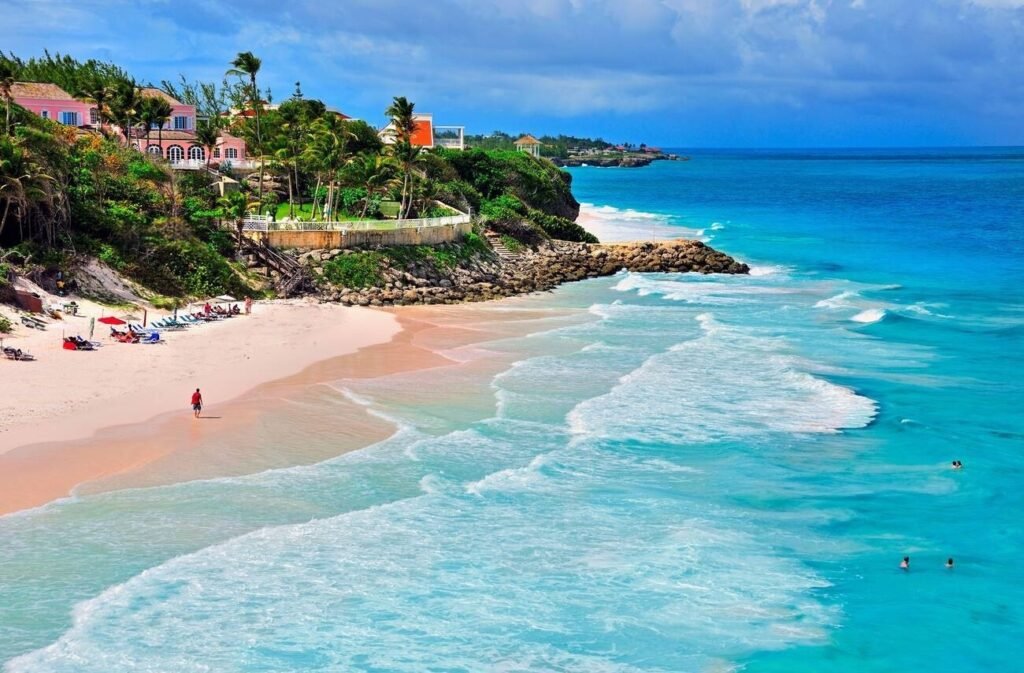An elevated view of the dramatic Crane Beach in Barbados, showing its pink-tinged sand, turquoise waves, and the historic Crane Resort on the cliffs.