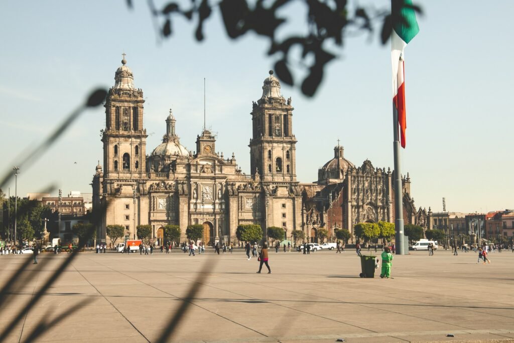 The Mexico City Metropolitan Cathedral viewed from the Zócalo, with a large Mexican flag waving nearby on a sunny day.