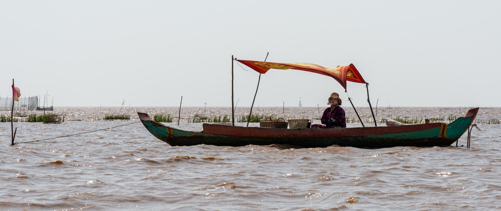 A person in a traditional wooden boat with a red and yellow canopy on the expansive Tonlé Sap Lake in Cambodia, reflecting the unique floating village life.