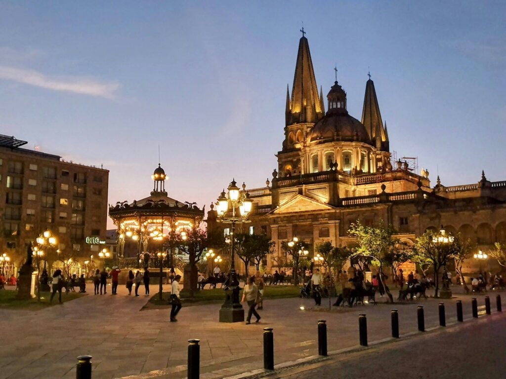 The Guadalajara Cathedral and a lighted gazebo in the Plaza de Armas at twilight, with people enjoying the evening.