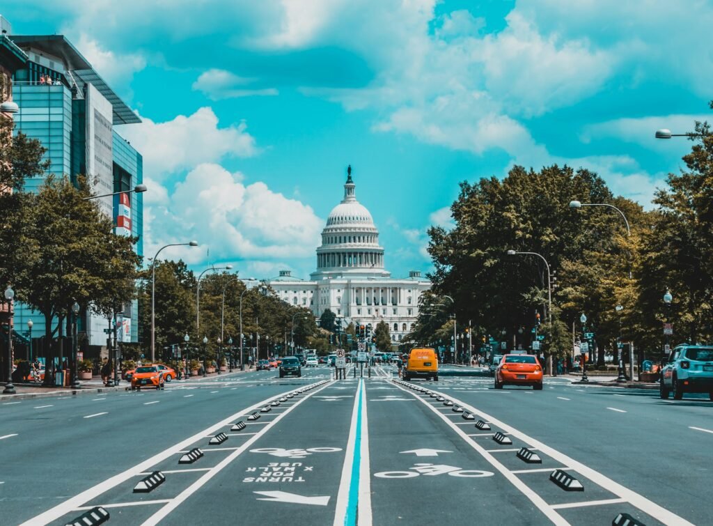A view down a wide avenue in Washington, D.C., leading to the iconic U.S. Capitol Building under a bright, cloudy sky.