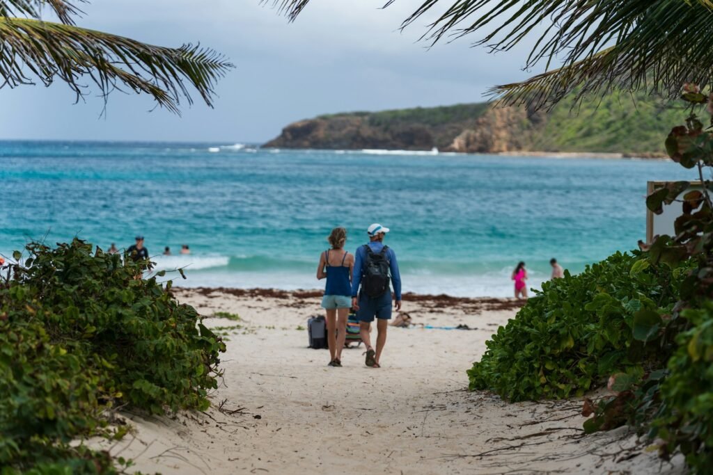A couple walking down a sandy path onto the white sands of Flamenco Beach in Culebra, Puerto Rico, with turquoise water in the background.