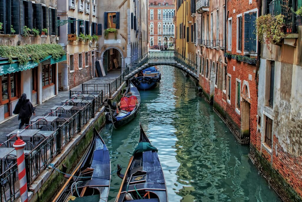 Top 10 European Cities : A quiet canal in Venice, Italy, with gondolas moored alongside a waterside cafe and a small bridge in the background.