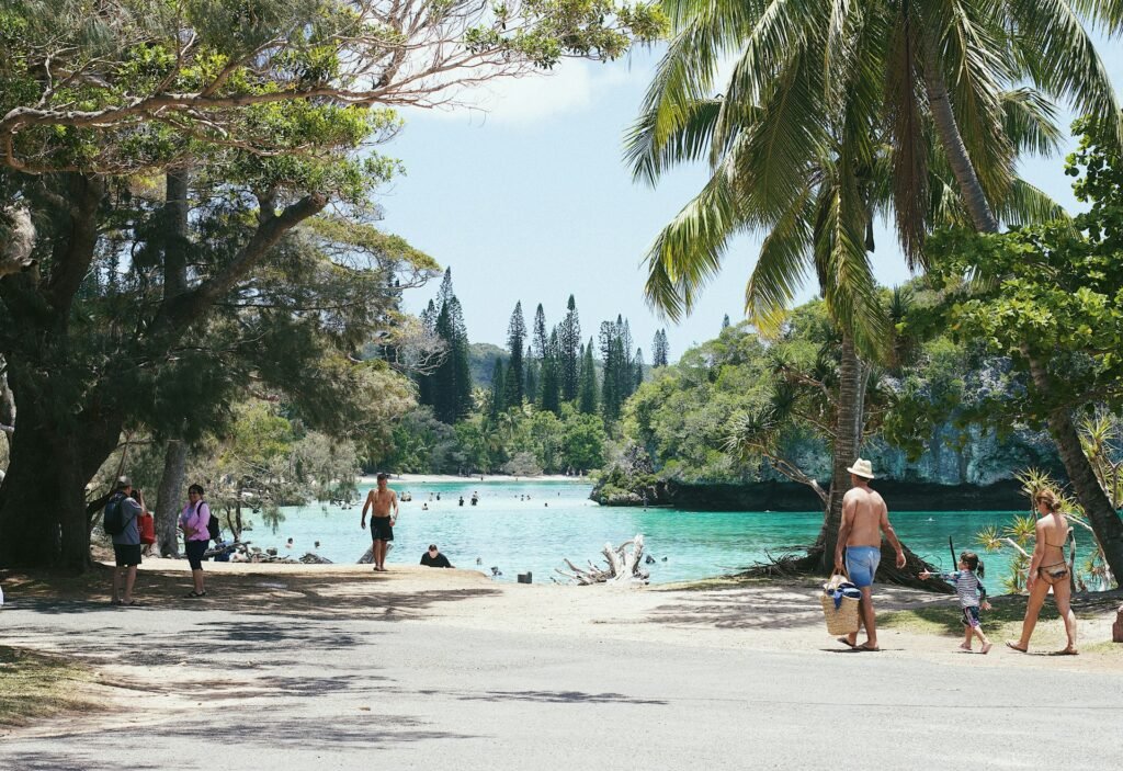 A family walking towards a beautiful natural swimming pool with turquoise water, surrounded by lush pine and palm trees in New Caledonia.