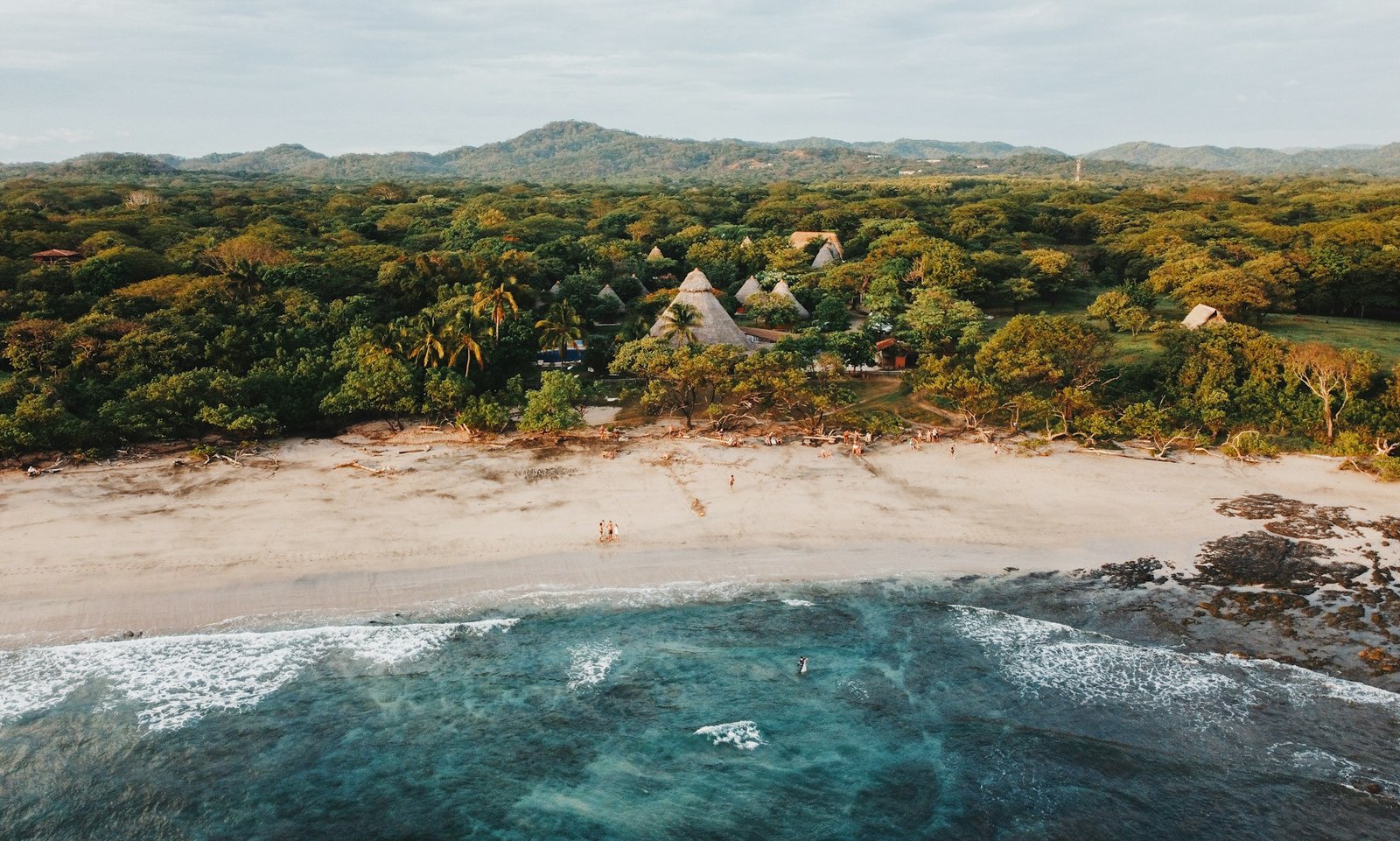 An aerial view of a secluded beach and eco-lodge in Central America, with turquoise water, a sandy shore, and a lush jungle backdrop.
