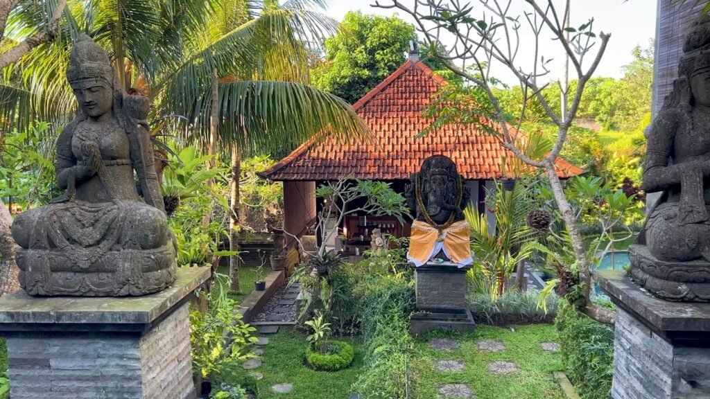 A tranquil garden in Ubud, Bali, featuring traditional stone statues, a Hindu shrine, and lush tropical plants.