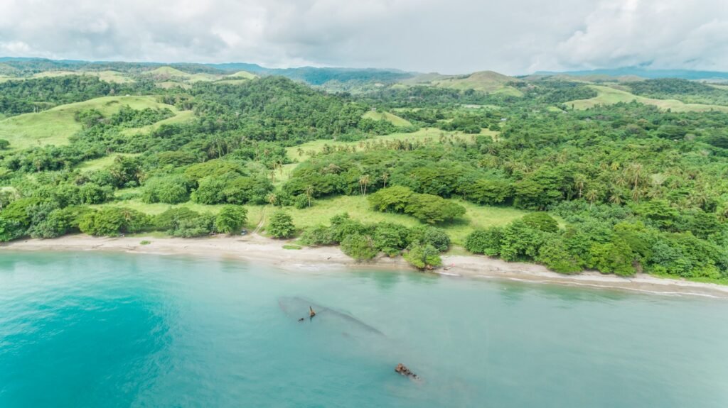 An aerial view of a sunken WWII shipwreck in the shallow turquoise water off the coast of the Solomon Islands, with lush green hills in the background Pacific islands to visit.