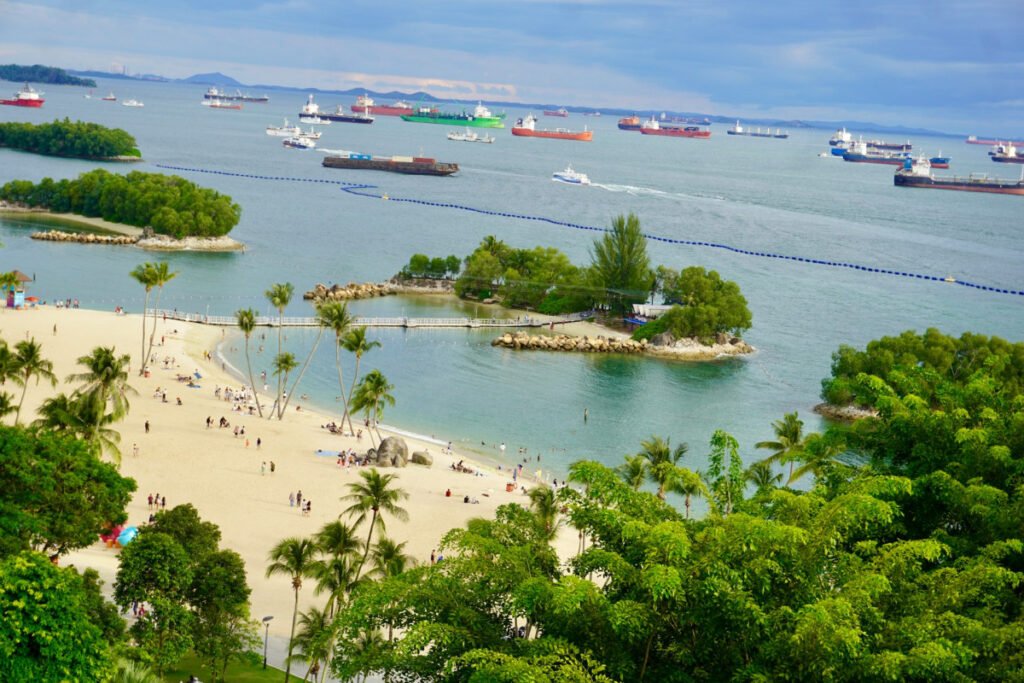 An elevated view of a sandy beach on Sentosa Island, Singapore, with palm trees, lush greenery, and cargo ships in the background.