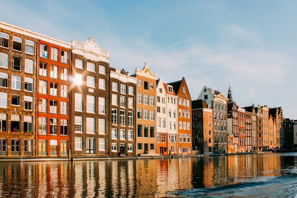 A row of colorful, historic Dutch canal houses reflecting in the water in Amsterdam, with the sun flaring off a window.