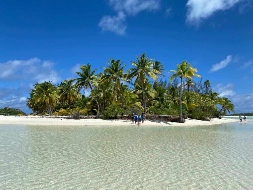 A family standing on the white sand beach of a small tropical island (motu) in the Cook Islands, surrounded by palm trees and clear, shallow water.