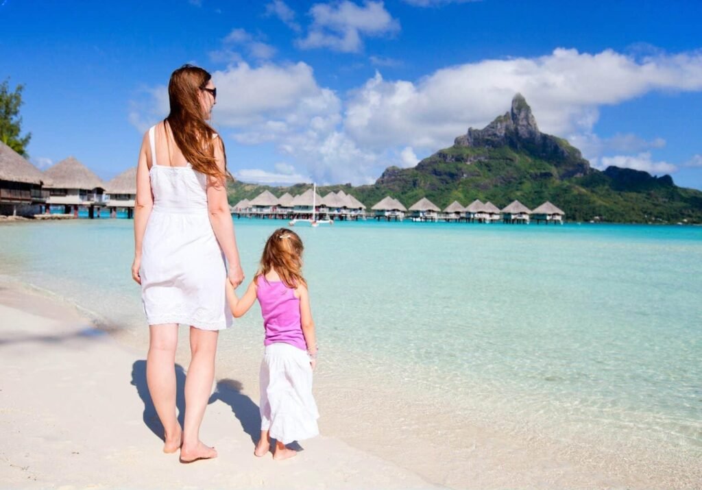 A mother and daughter holding hands on a beach in Bora Bora, looking at overwater bungalows with Mount Otemanu in the background.