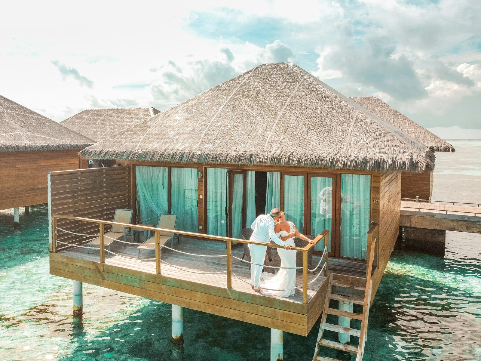 A couple dressed in white kissing on the deck of a luxurious overwater bungalow with a thatched roof, surrounded by turquoise water.