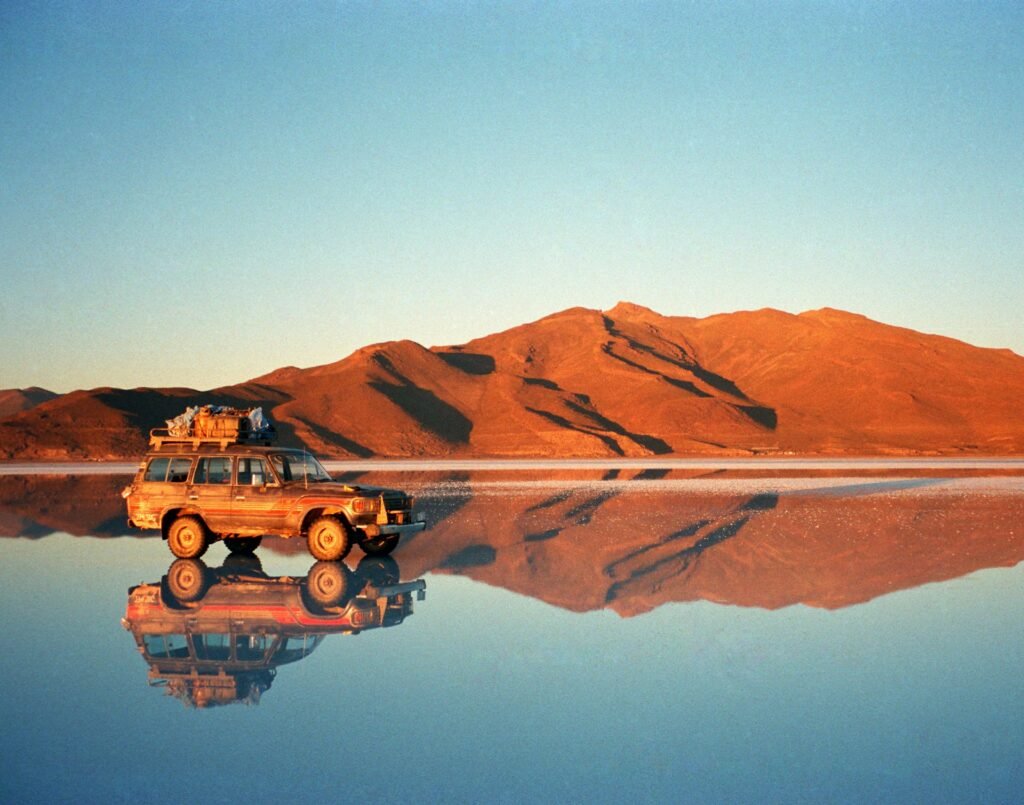 A 4x4 vehicle on the Salar de Uyuni salt flats in Bolivia at sunset, with a perfect mirror reflection in the water.