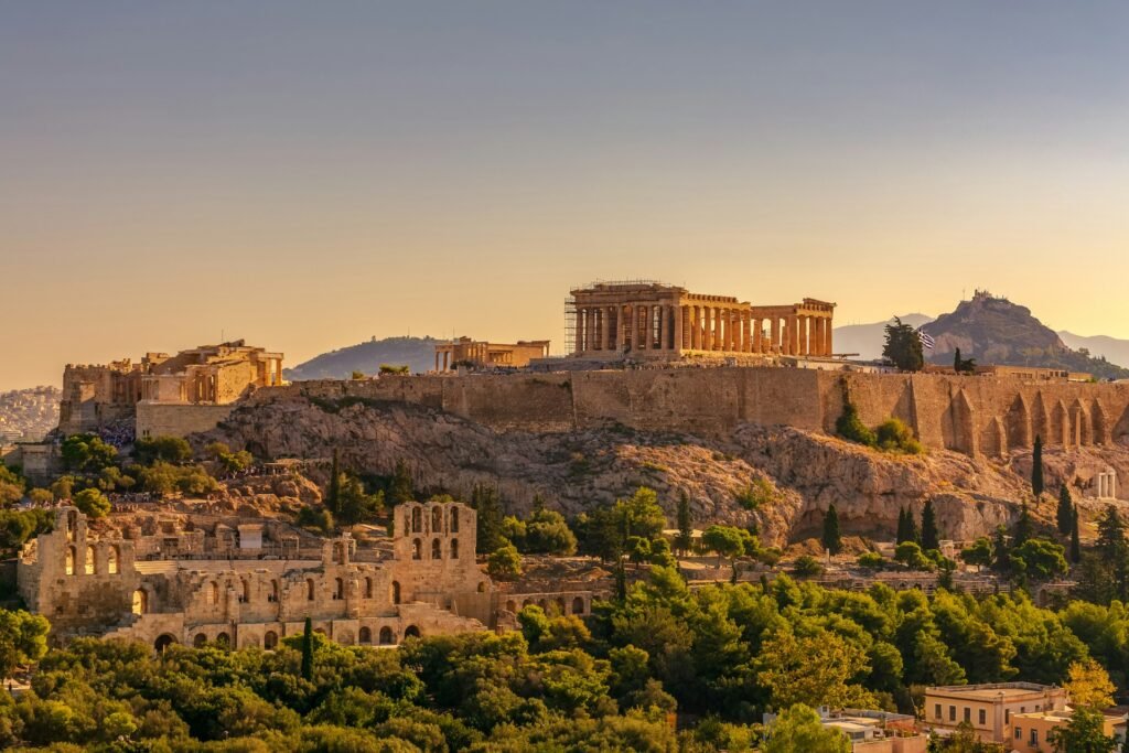 The ancient Acropolis of Athens, including the Parthenon, illuminated by the golden light of sunset, with Mount Lycabettus in the background.