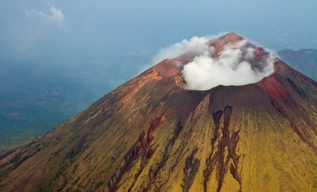 An aerial view of the active Cerro Negro volcano in Nicaragua, with steam rising from its crater and dark volcanic ash covering its steep slopes.