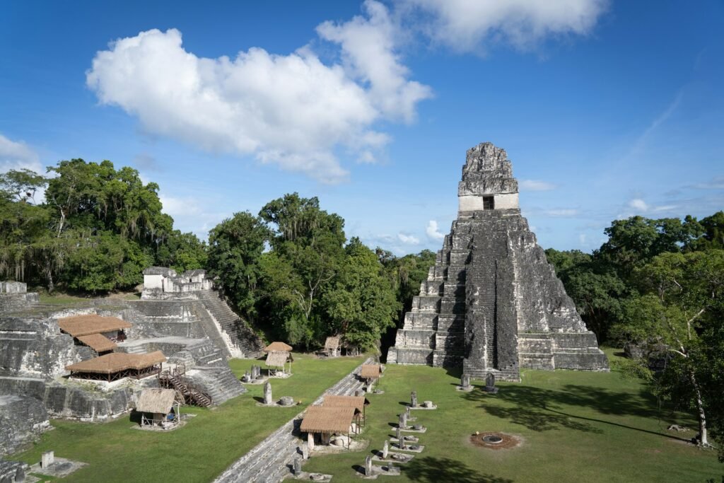 A wide view of the Great Plaza in Tikal, Guatemala, showing the iconic Temple I and the North Acropolis rising above the jungle canopy under a blue sky.