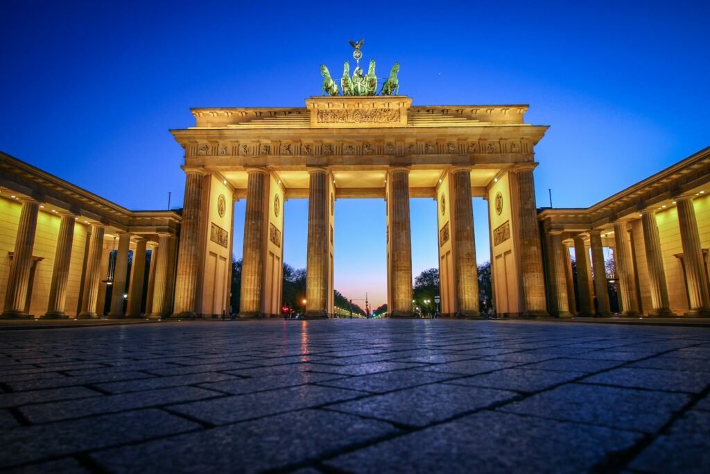 A low-angle view of the illuminated Brandenburg Gate in Berlin, Germany, at twilight, with the cobblestone plaza in the foreground.