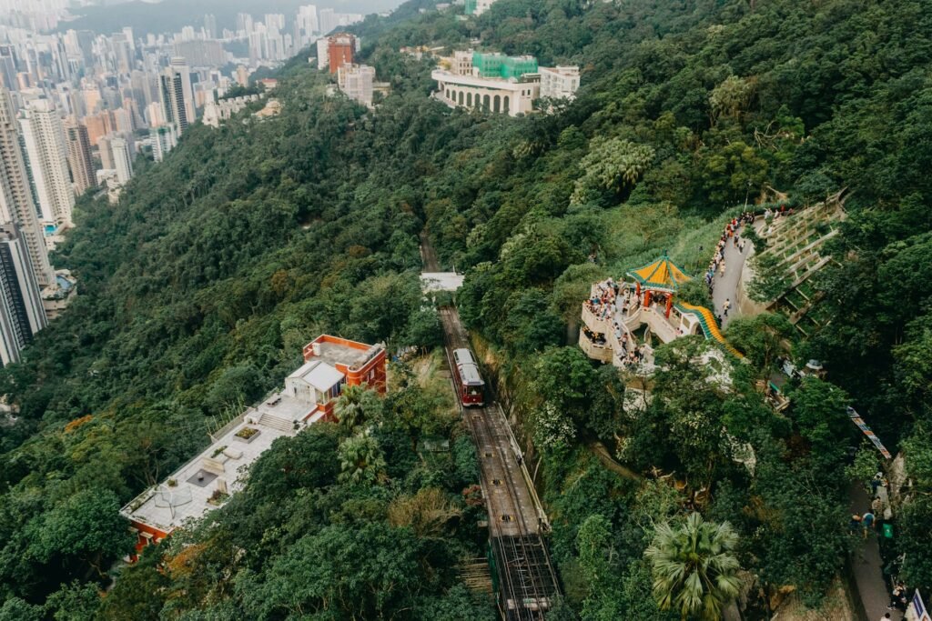 An aerial view of the historic red Peak Tram ascending the lush, green Victoria Peak in Hong Kong, with the city skyline in the background.