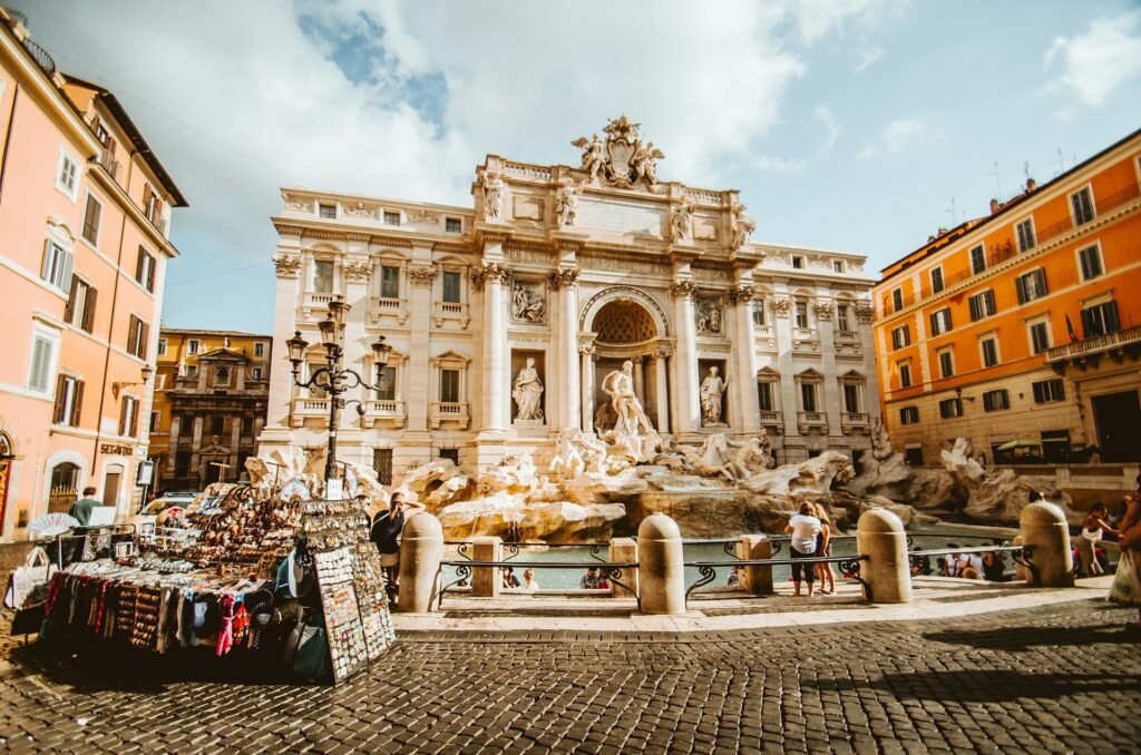 The magnificent Trevi Fountain in Rome on a sunny day, with tourists and a souvenir stand in the plaza.