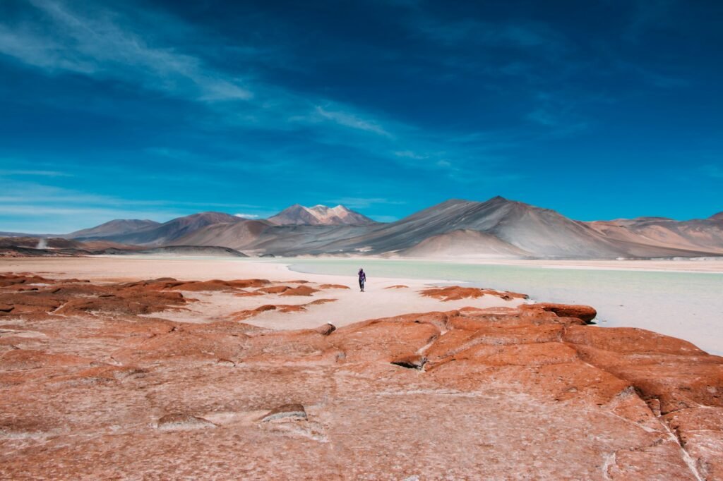 A lone person walking in the otherworldly landscape of the Atacama Desert in Chile, with red rocks, a salt lagoon, and mountains.
