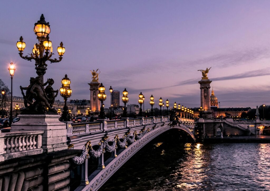 The ornate Pont Alexandre III bridge in Paris, illuminated by glowing street lamps as it crosses the River Seine at twilight.