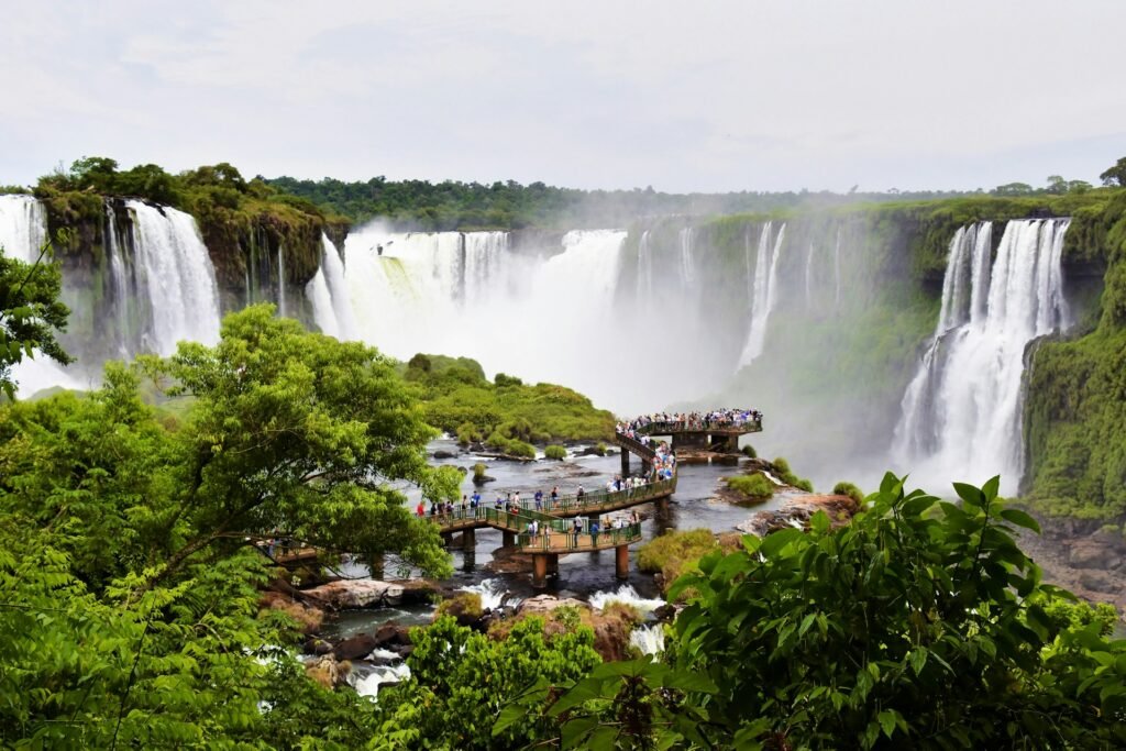 A panoramic view of the powerful Iguazu Falls in Argentina, with tourists on a viewing platform surrounded by lush rainforest.